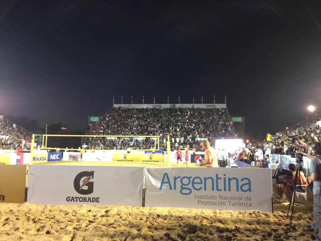 A full beach volleyball stadium at night, seen from courtside