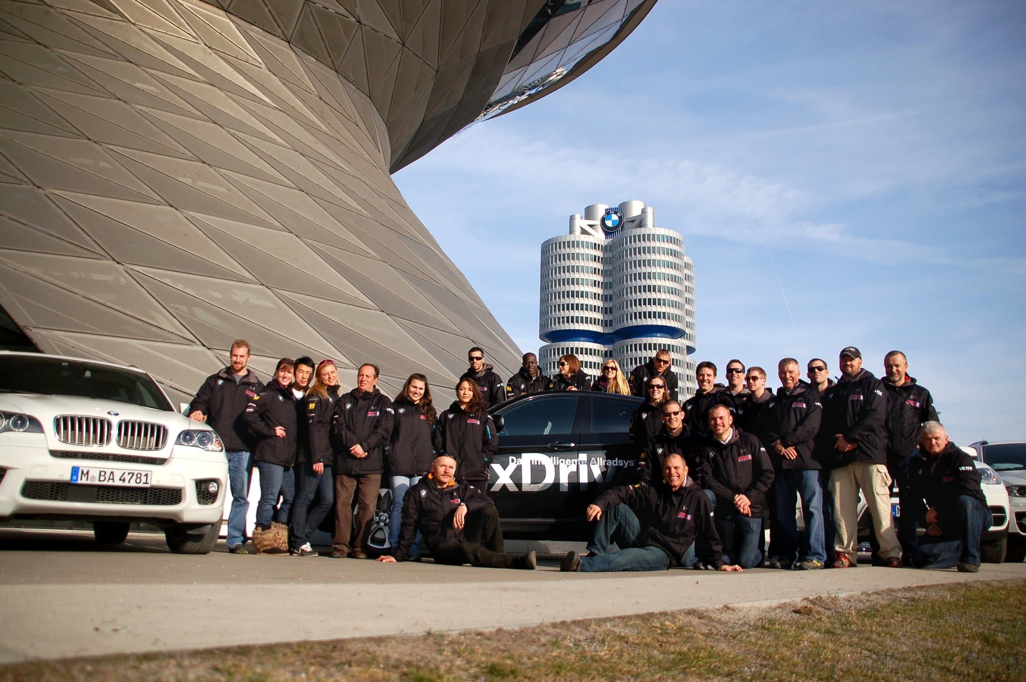 The USA Skeleton team group in matching dark jackets posing beside BMW cars in front of the BMW headquarters tower in Munich