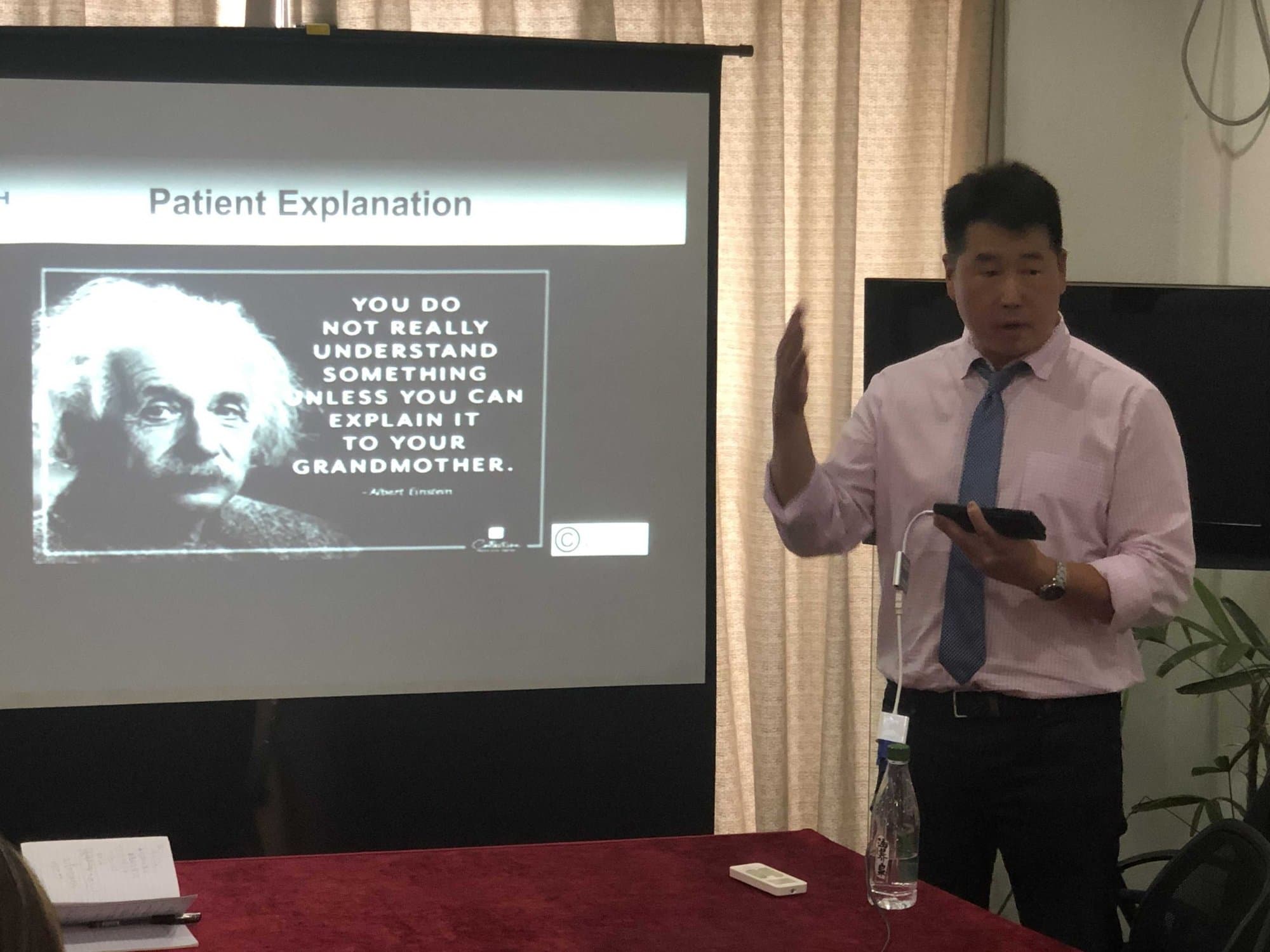 Dr. Koh in a shirt and tie, lecturing beside a projector screen showing a slide titled Patient Explanation