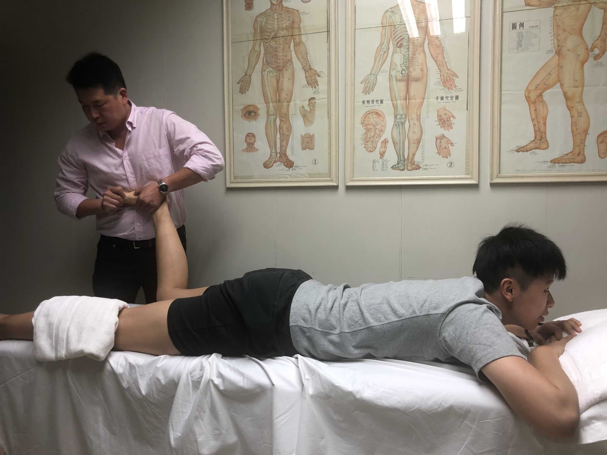 Dr. Koh treating the leg of an athlete lying face down on a treatment table, with anatomy charts on the wall