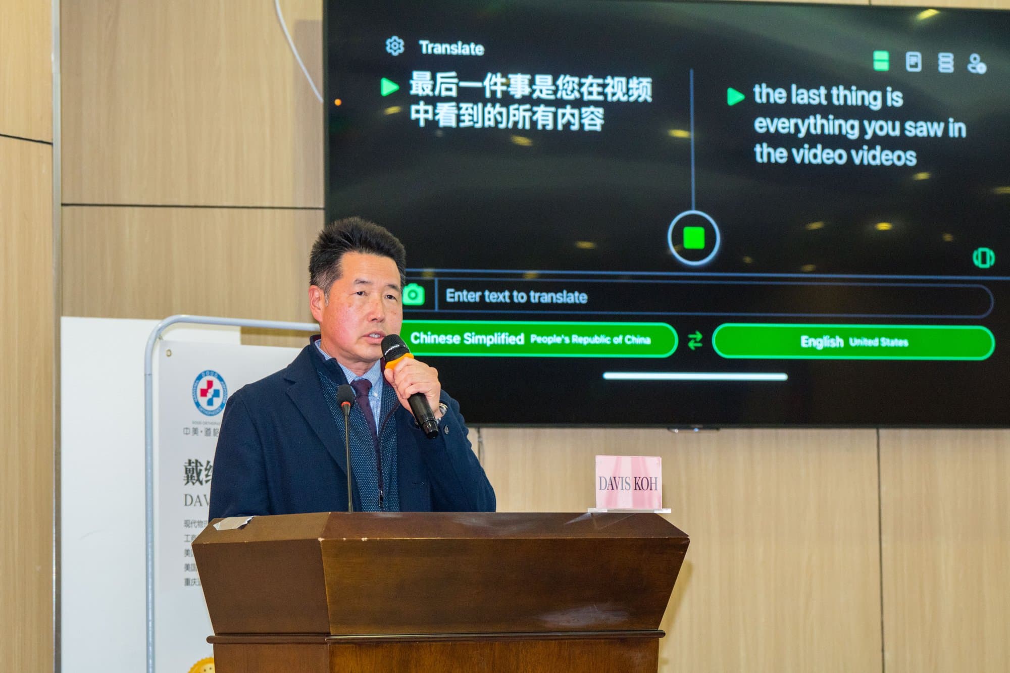 Dr. Koh speaking into a microphone at a wooden lectern with a live translation screen behind him
