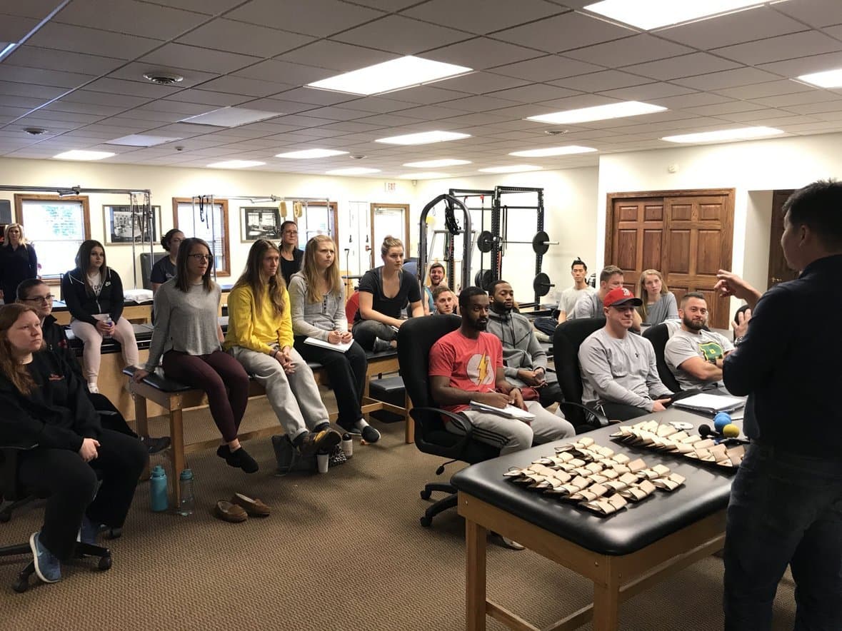 Course attendees seated around treatment tables set with rows of instruments, listening to Dr. Koh