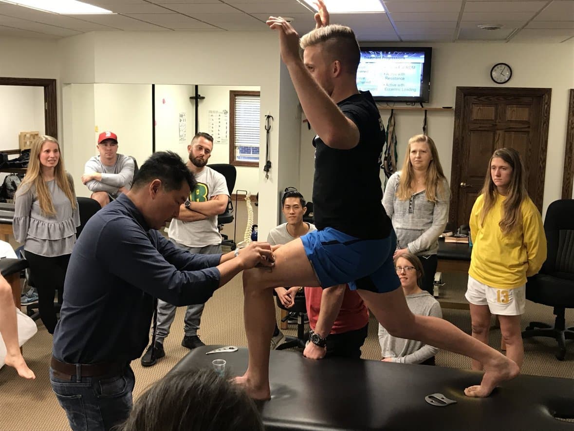 Dr. Koh kneeling to check an attendee's knee position during a lunge while the class watches