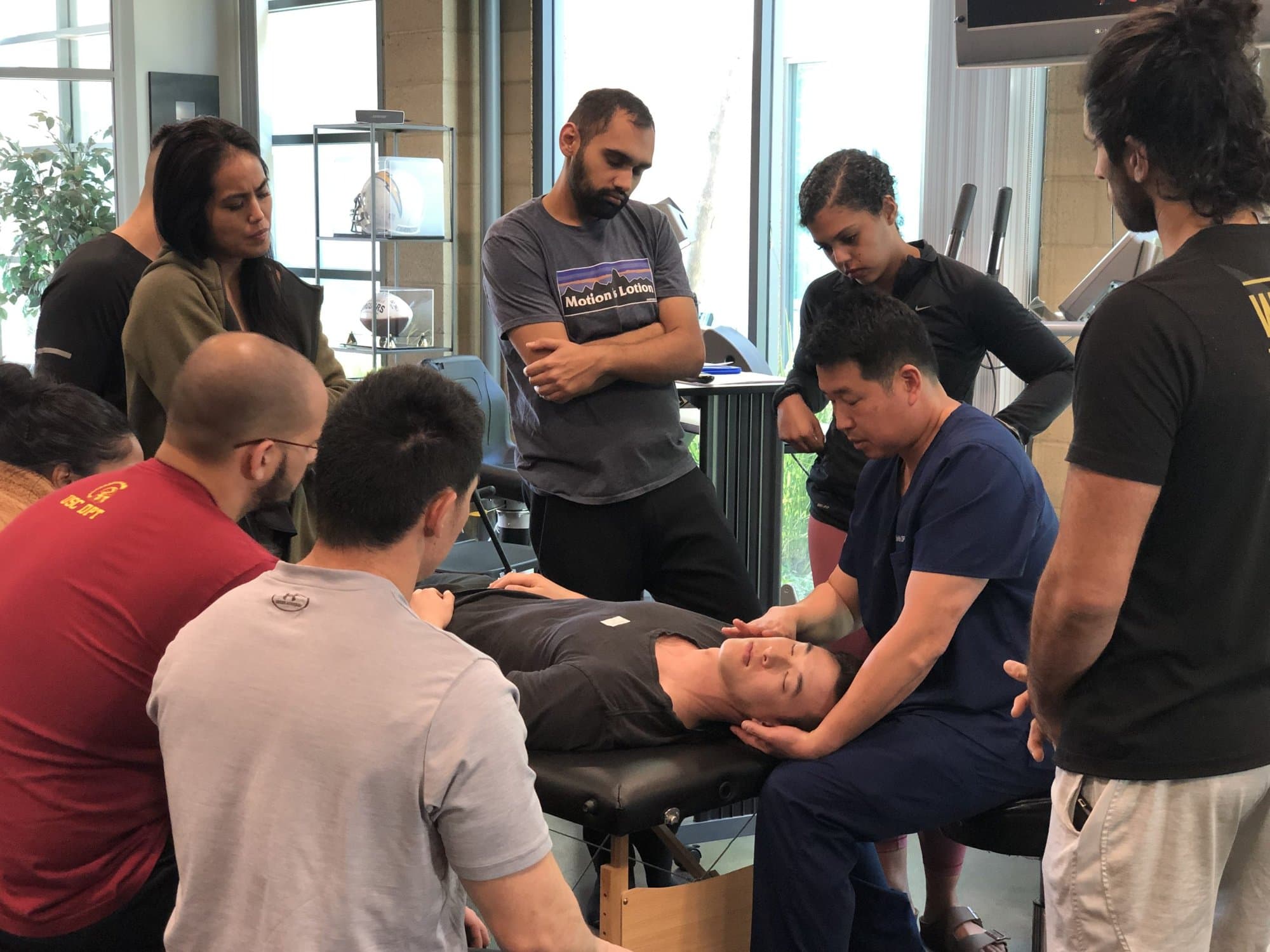Dr. Koh demonstrating a neck technique on an attendee lying on a table, with other attendees gathered around