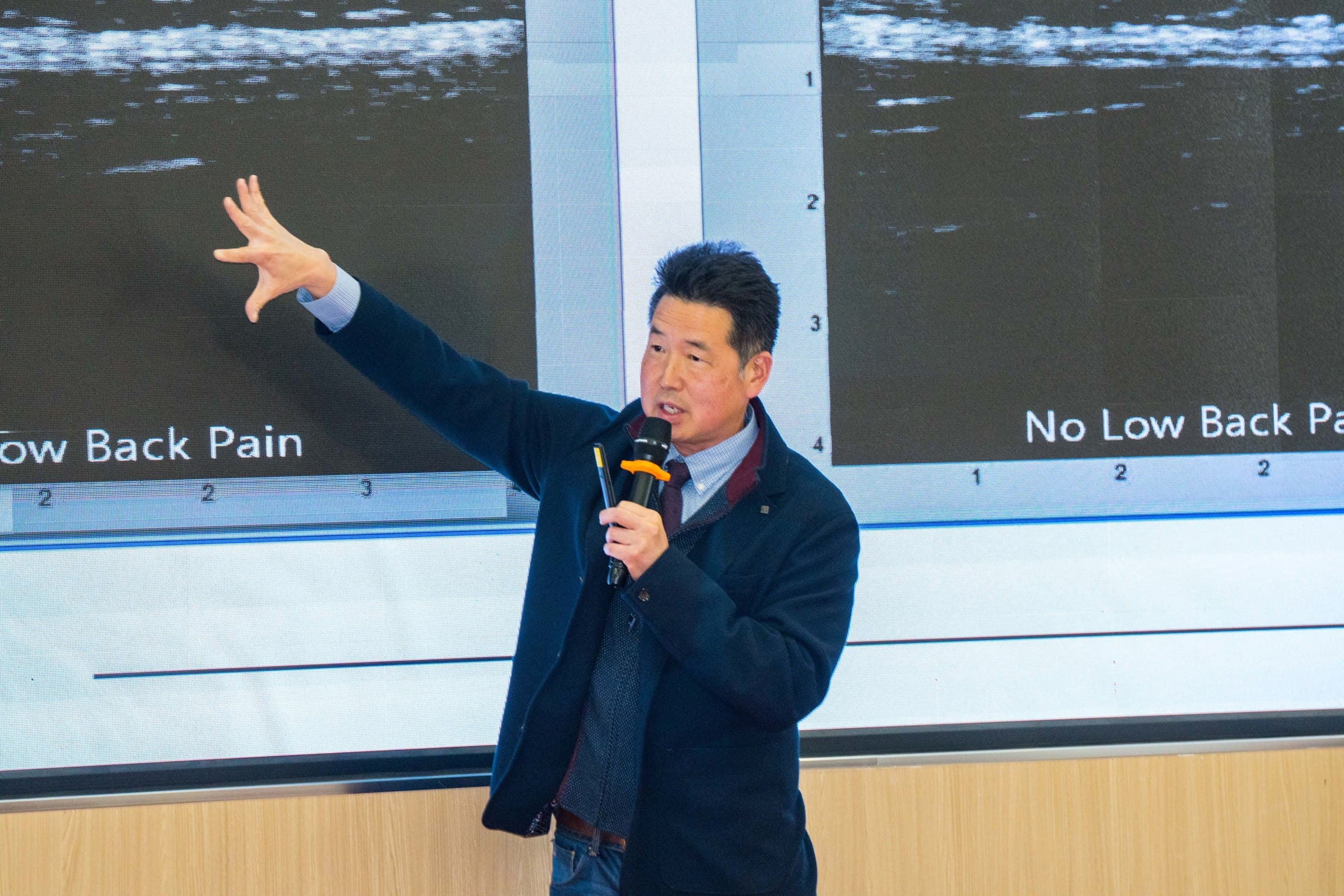 Dr. Koh holding a microphone and gesturing toward ultrasound images on a large screen during a lecture on low back pain
