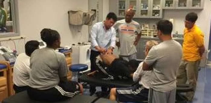 Dr. Koh demonstrating a technique on a person lying on a treatment table while staff members gather around and watch