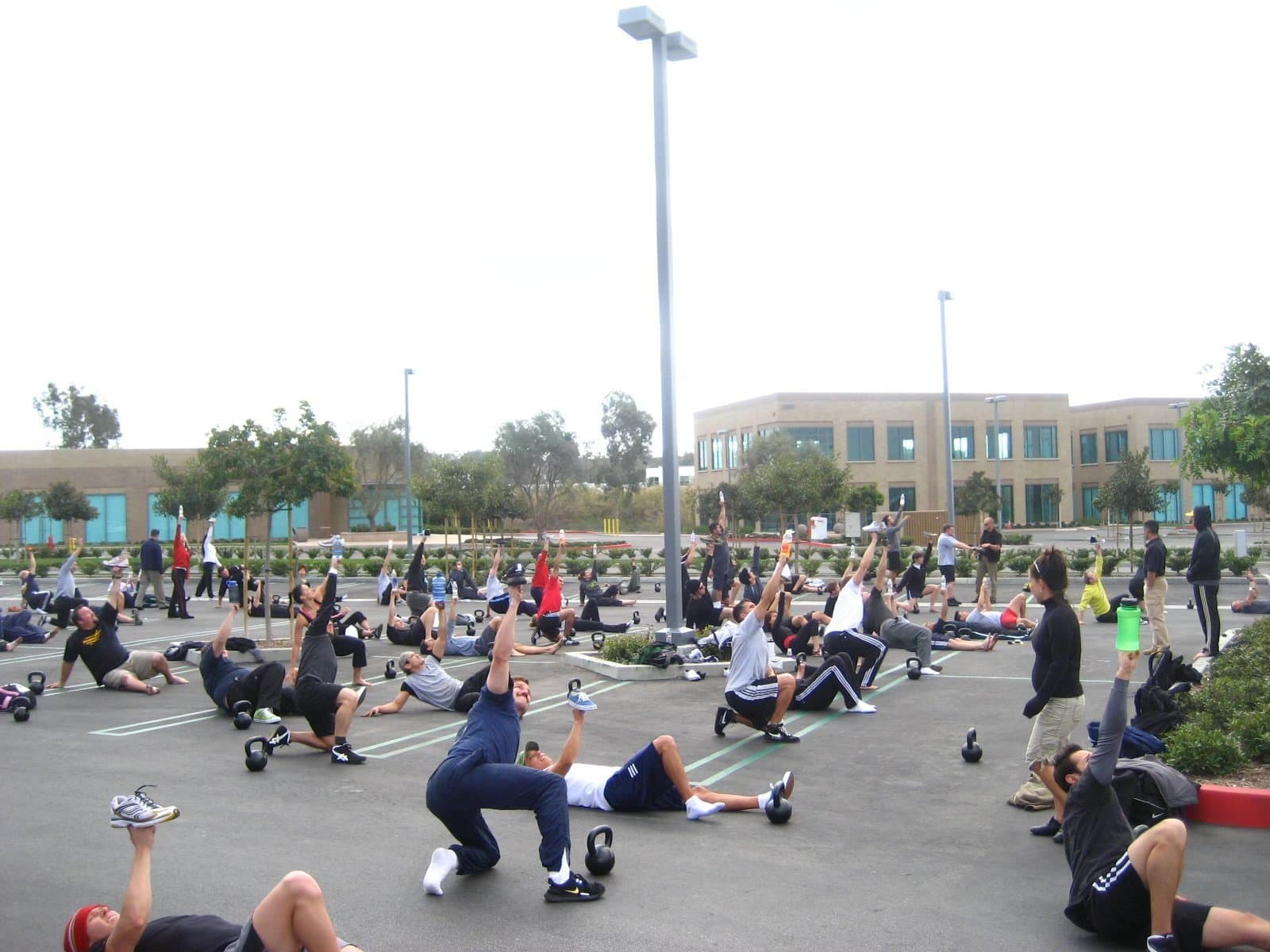A large group practicing kettlebell exercises outdoors in a parking area