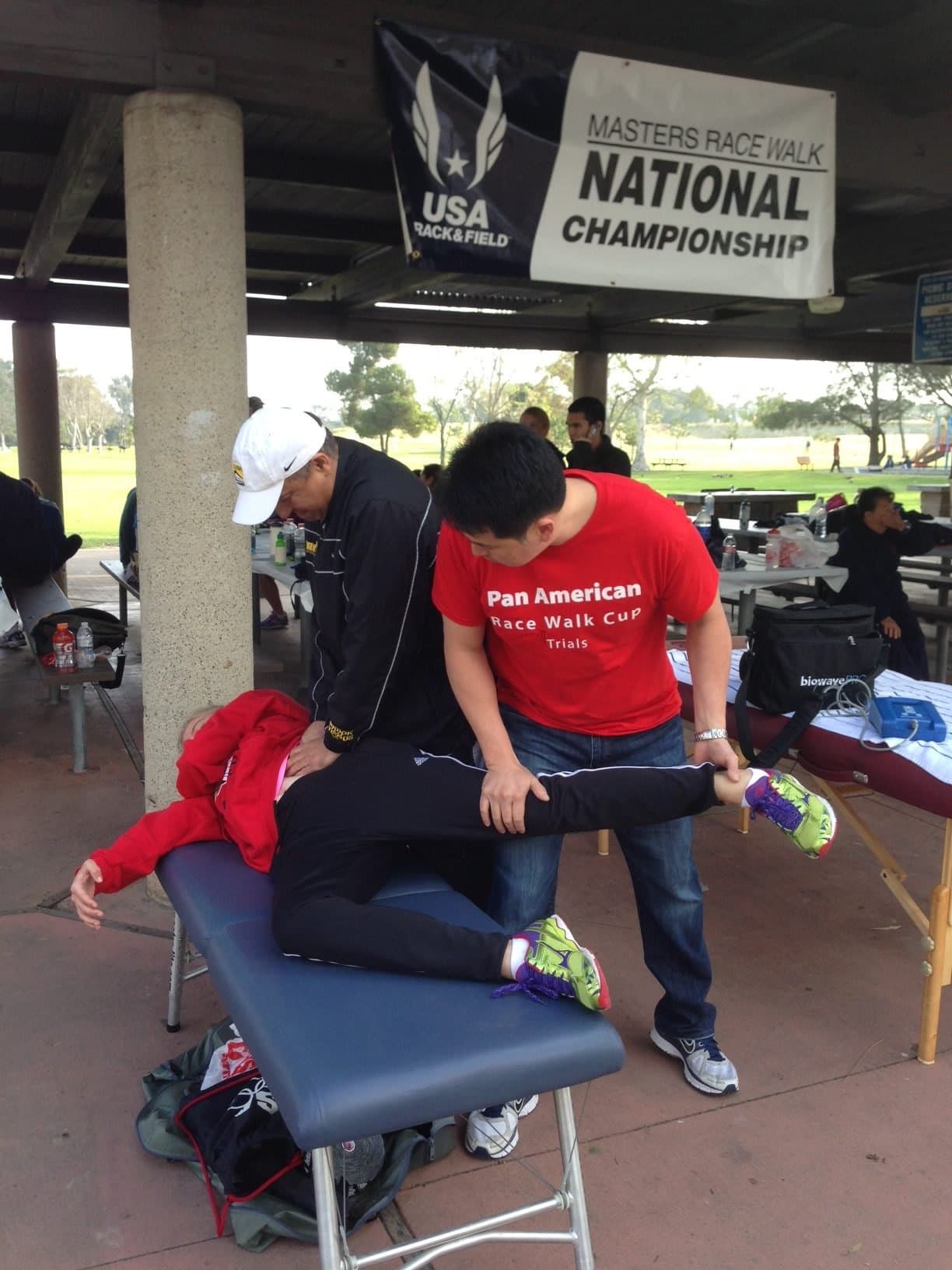 Dr. Koh and another clinician treating an athlete lying face down on a portable table, under a USA Track and Field Masters Race Walk National Championship banner