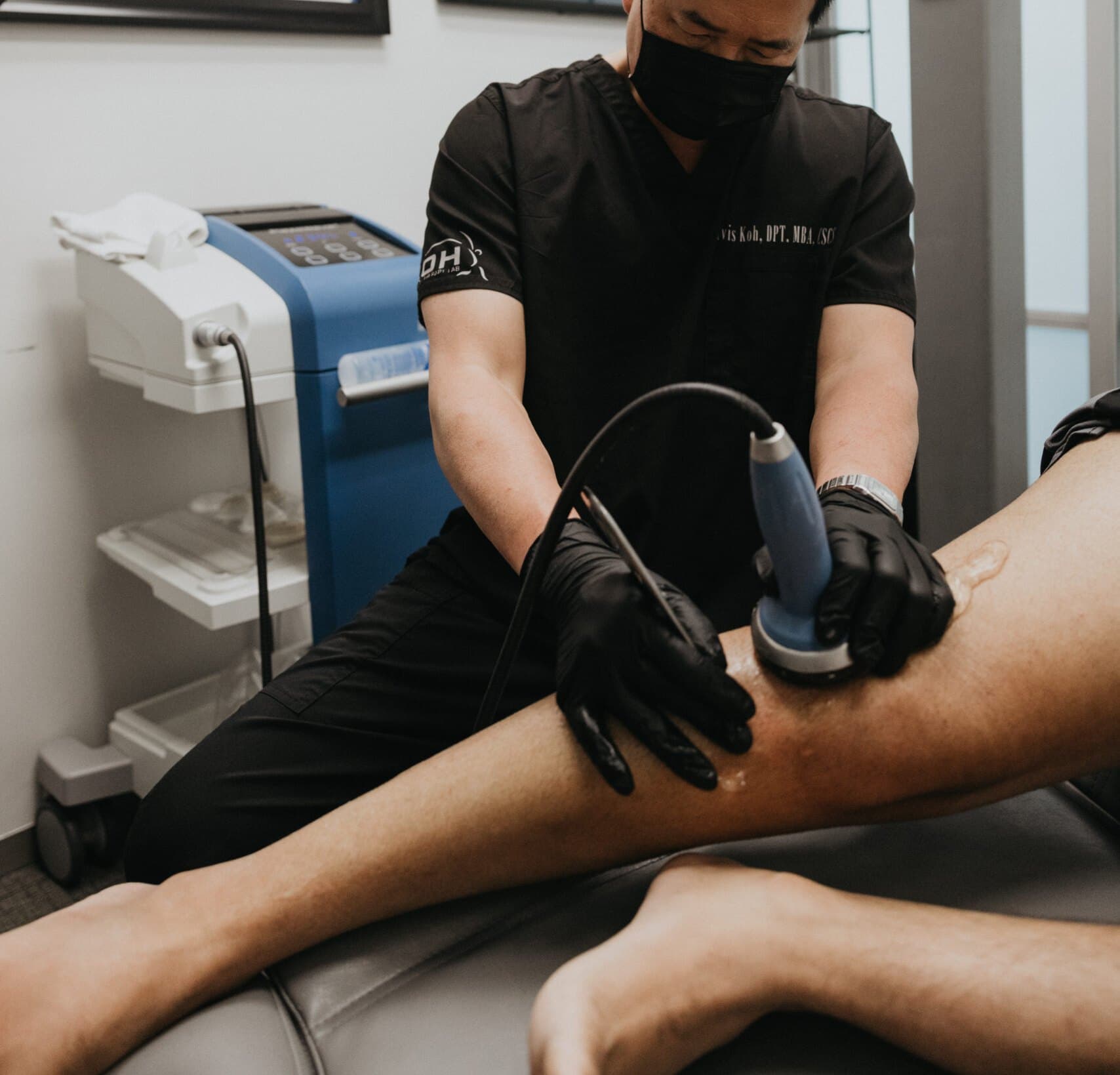 Dr. Koh, wearing a mask and gloves, applying a shockwave therapy device to a client's lower leg in a private treatment room