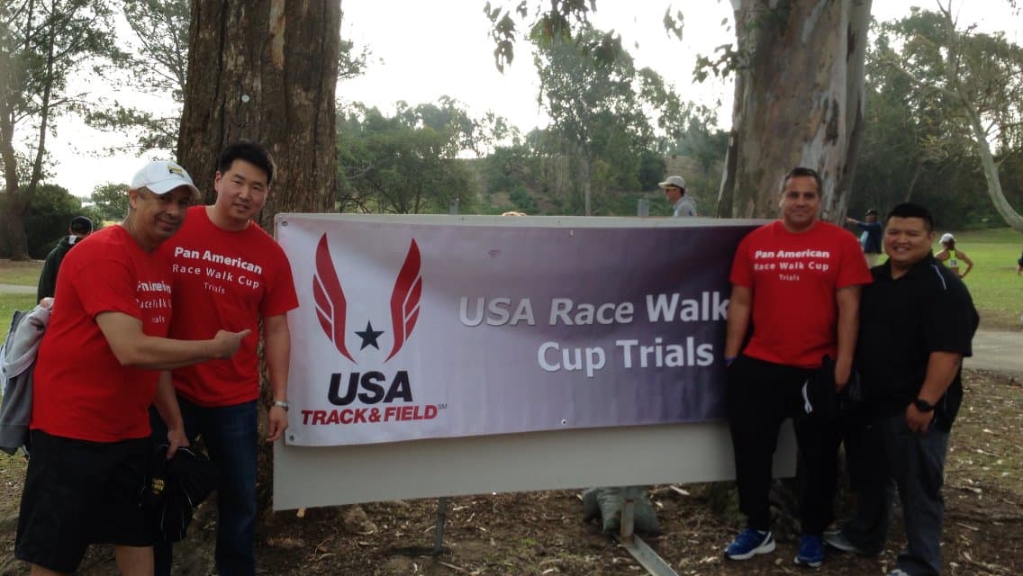 Dr. Koh and colleagues in red Pan American Race Walk Cup shirts beside a USA Track and Field Race Walk Cup Trials banner