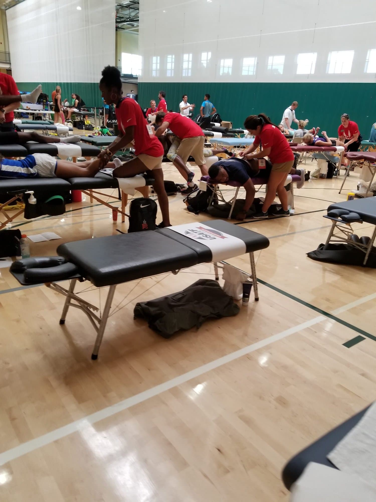A gymnasium set up as an athlete treatment area, with clinicians in red shirts working at rows of portable tables