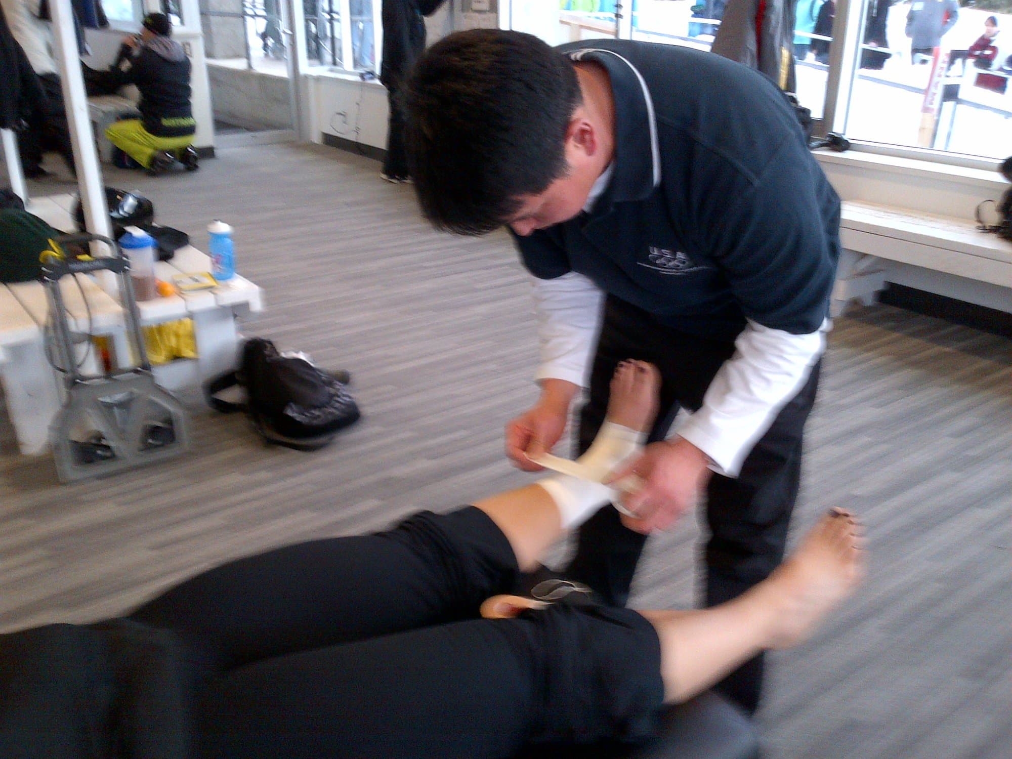 Dr. Koh bending to tape the ankle of an athlete lying on the floor of a team treatment area