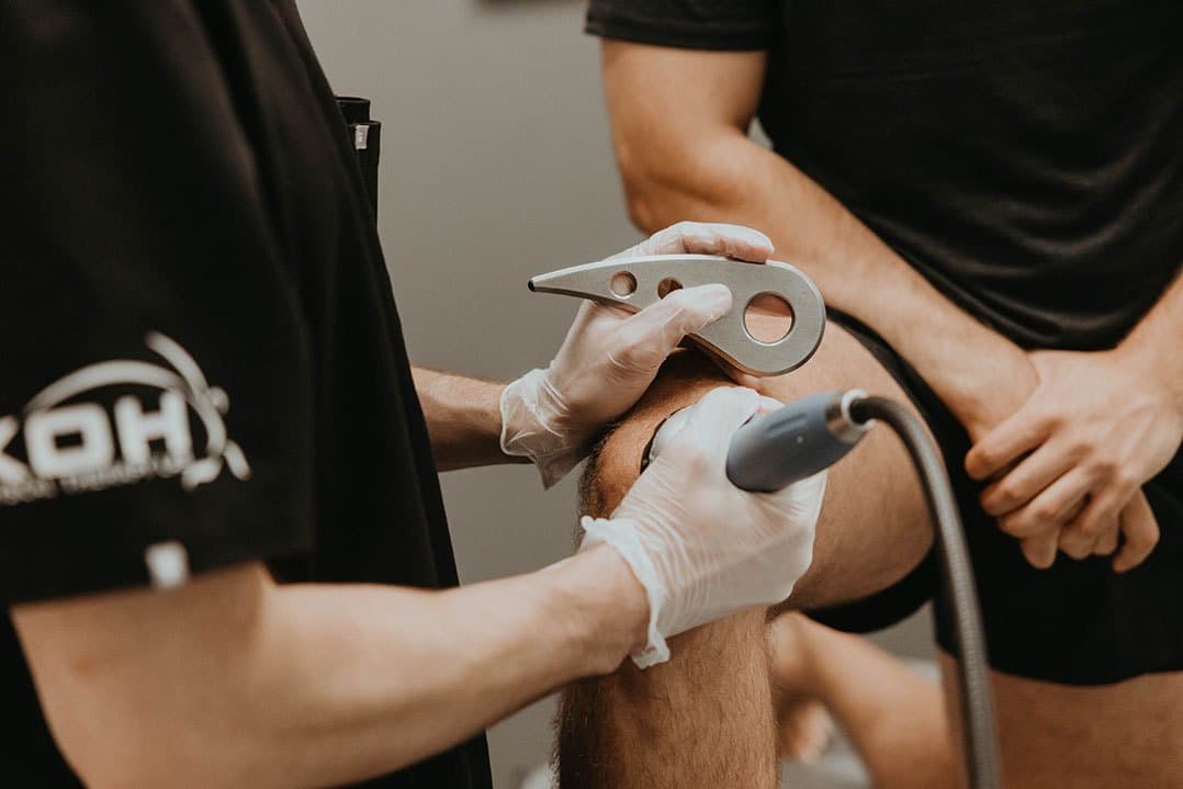 Gloved hands holding a steel soft tissue instrument and a treatment applicator against a client's knee