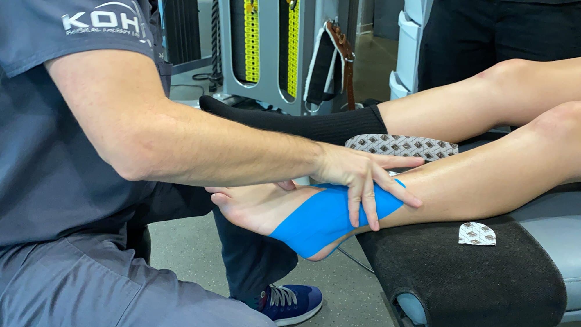 A clinician's hands applying blue kinesiology tape to a client's ankle on a treatment table